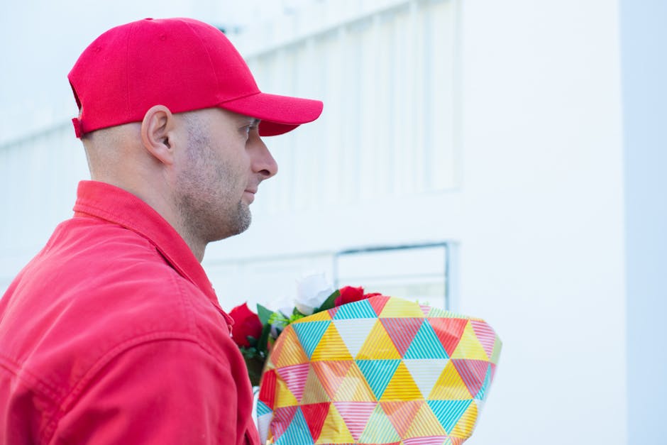 A man wearing a red cap and matching red jacket is holding a colorful, geometrically patterned gift box with a floral bouquet inside, featuring red, white, and green flowers. The bouquet is wrapped in