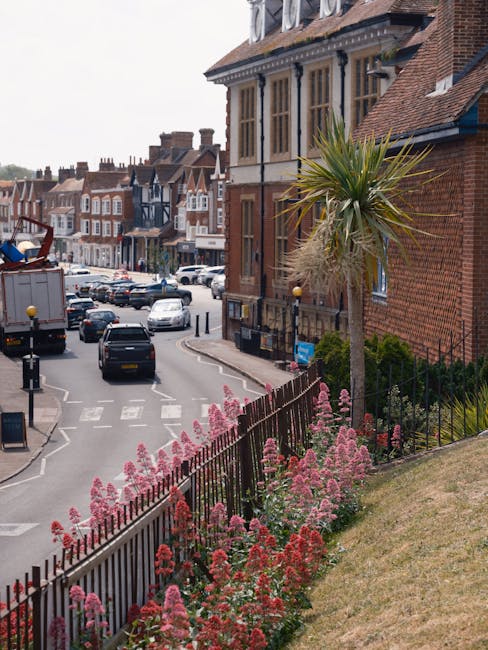 A floral display along a street in Chertsey featuring a row of pink and red flowering plants in the foreground, planted behind a black iron fence on a grassy slope. In the background, a tall, slender