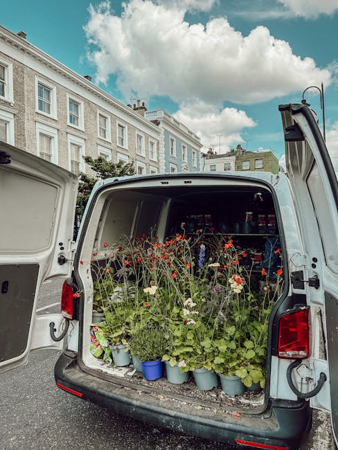 The open back of a white delivery van filled with potted plants and fresh flowers, including red poppies, white daisies, and green foliage, placed on the vehicle's floor in light blue pots and plastic