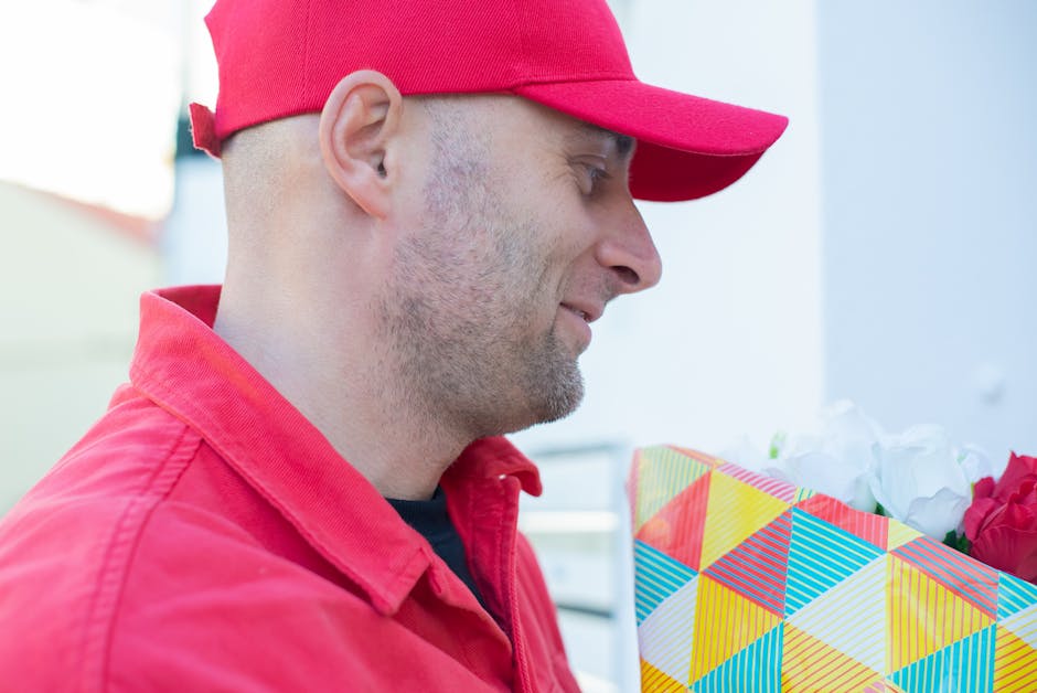 A delivery man dressed in a red uniform and matching red cap is holding a floral gift wrapped in decorative colorful geometric-patterned paper, containing a bouquet of white and red flowers with green