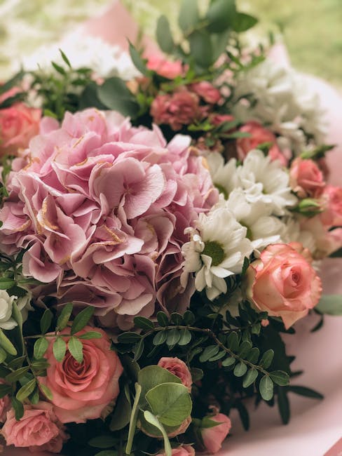 A close-up of a floral arrangement featuring large, soft pink hydrangea blossoms with delicate, ruffled petals, complemented by small, pale peach roses and white daisies with yellow centers. The arran