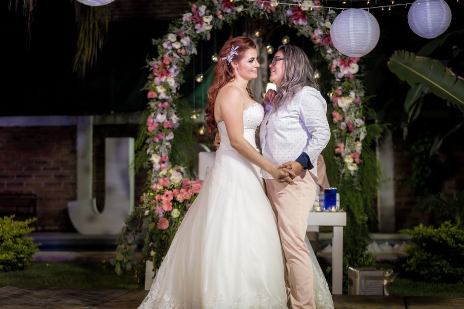 A bride with long red hair styled in loose waves, wearing a strapless white wedding gown with delicate lace detailing at the hem, smiles warmly while holding hands with a woman dressed in a light gray