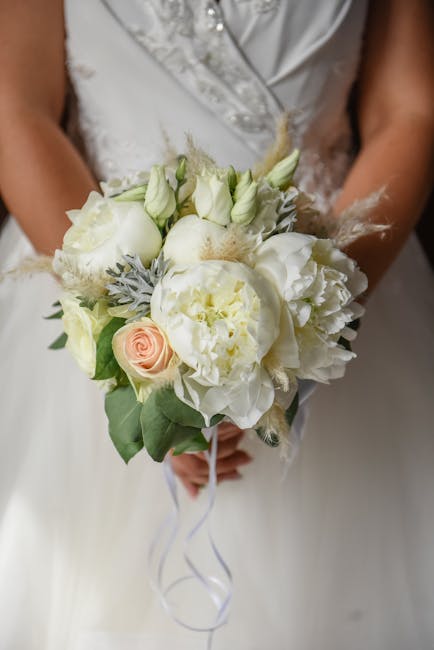 A bride holds a wedding bouquet featuring an arrangement of fresh, creamy white flowers including roses, peonies, and lisianthus, accented with soft gray foliage and delicate feathers. The bouquet is 