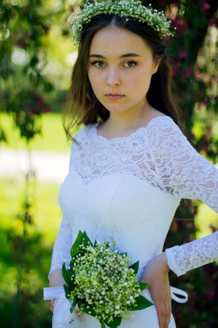 A young woman dressed in a white lace wedding dress holds a small bouquet of fresh lily of the valley flowers, featuring tiny white bell-shaped blooms and green leaves. She wears a delicate floral cro