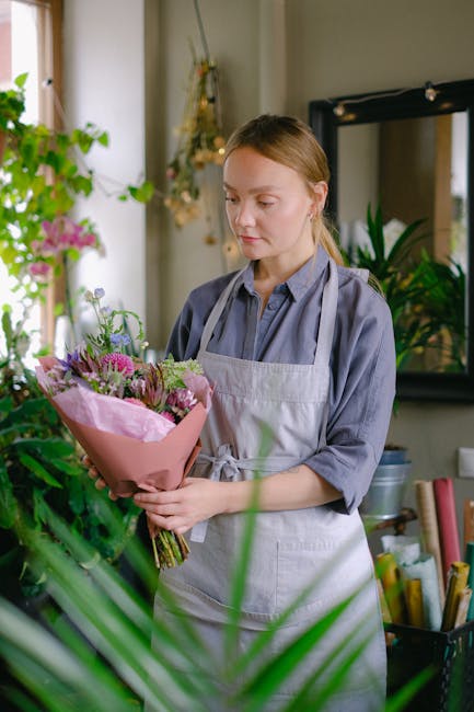 A young woman with fair skin and light brown hair, tied back, is standing indoors in a floral shop or studio environment, gently holding a bouquet of fresh flowers. The bouquet features a variety of b