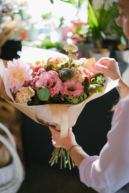 A person holding a large, fresh bouquet of mixed flowers, including pink peonies, soft white roses, and delicate purple blossoms, all arranged in a natural, rounded style. The bouquet is wrapped in cr